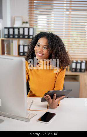 Eine attraktive, lächelnde schwarze Geschäftsfrau, die an ihrem Schreibtisch im Büro arbeitet und ihr digitales Tablet hält, während sie auf den Computerbildschirm blickt. Stockfoto