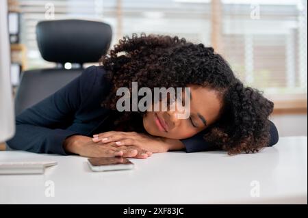 Eine müde, verschlafene schwarze Geschäftsfrau, die ein Nickerchen an ihrem Schreibtisch im Büro macht, während der Arbeit schläft und nachmittags eine Pause einlegt. Stockfoto