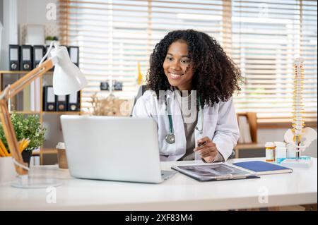 Eine selbstbewusste, professionelle schwarze Ärztin in einem weißen Kleid arbeitet an ihrem Laptop-Computer an ihrem Schreibtisch im Büro und liest online medizinische Koffer Stockfoto