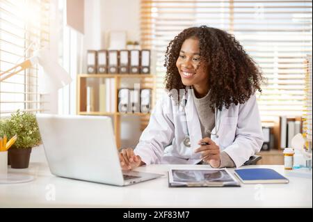 Eine selbstbewusste, professionelle schwarze Ärztin in einem weißen Kleid arbeitet an ihrem Laptop-Computer an ihrem Schreibtisch im Büro und liest online medizinische Koffer Stockfoto