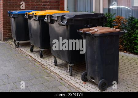 Eine Reihe von vier Mülltonnen mit Rädern, jeder mit einem anderen farbigen Deckel vor einem Gebäude. Stockfoto