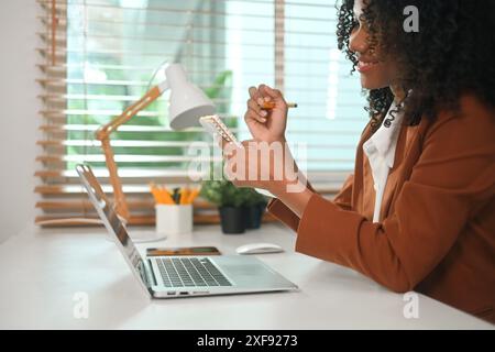 Seitenansicht einer lächelnden afroamerikanischen Geschäftsfrau mit Laptop und Notizen in einem Notizbuch Stockfoto