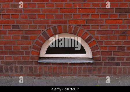 Eine Backsteinmauer mit einem bogenförmigen Fenster, mit einem weißen Rahmen und einem dunklen, undurchsichtigen Glas. Stockfoto