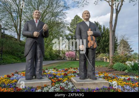 Statuen von zwei Männern, einer mit Geige, der andere mit Dirigentenstab, stehen im Frühling in einem bunten Garten, Skulpturen in Siegen Stockfoto