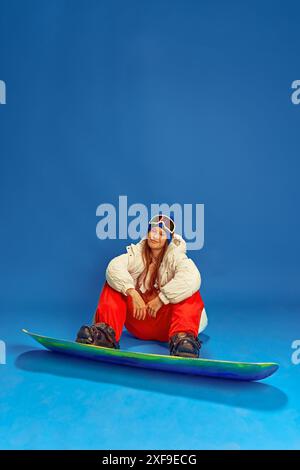 Frau, die Sommer- und Winterkleidung kombiniert, sich mit ihrem Snowboard entspannt und vor blauem Studiohintergrund sitzt. Extreme-Sport-Werbung Stockfoto