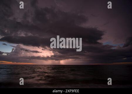 Gewitter dunkle Wolken mit Blitz und Donner über abendlichen Meerwasserwellen, die am Sandstrand zerbrechen. Meereslandschaft bei Sonnenuntergang Stockfoto