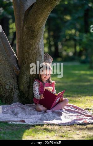 Ein kleines, süßes Mädchen sitzt neben einem großen Baum im Park. Er liest ein Buch mit Interesse, ein kleiner Teddybär sitzt in seinen Armen. Stockfoto