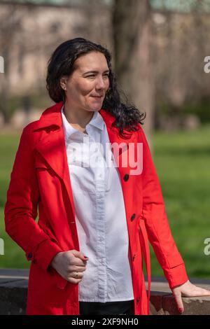 Porträt einer Frau mit dunklem Haar in rotem Regenmantel und Geschäftsbluse. Er steht unter freiem Himmel und blickt nachdenklich in die Ferne. Stockfoto