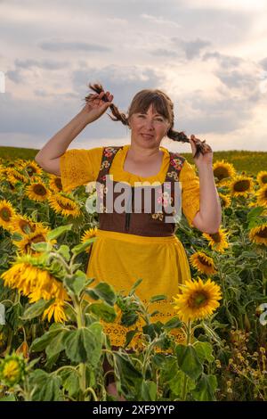 Eine Frau steht mitten auf einem Feld mit vielen gelben Sonnenblumen. Geflochtene Haare, gekleidet in deutscher Nationalkleidung, gelbe Bluse, brauner Sundr Stockfoto