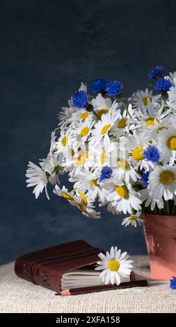 Ein sehr großer Strauß weißer Gänseblümchen und blauer Kornblumen in einem rosafarbenen Eimer, neben einem Buch auf einem Ledereinband. Wunderschönes Stillleben. Postkarte, congra Stockfoto