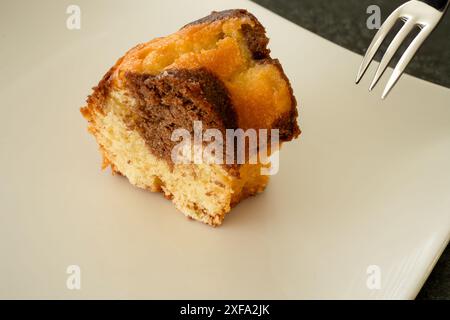 Ein Stück Marmorkuchen auf einem weißen Teller. Reflexion des Kuchens auf dem Teller. Schwarzer Hintergrund. Eine Gabel im Hintergrund von jemandem, der ein Stück davon will. Stockfoto