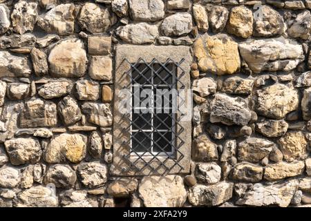 Textur der alten Steinmauer mit großen Kalksteinblöcken in verschiedenen Pastellfarben. In der Wand ein altmodisches Gitterfenster mit einem Metallgitter mit Stockfoto