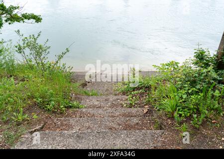 Schmale Treppe, die durch grüne Bürste in das Wasser eines Flusses führt. Tote Blätter auf dem Weg. Tageslicht und Wolken reflektieren auf dem Wasser. Stockfoto