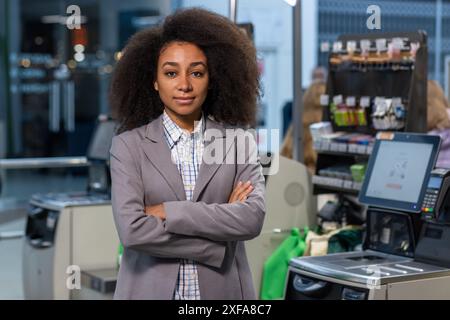 Ein selbstbewusster Kassierer im Supermarkt mit lockigen Haaren in Geschäftskleidung, der mit überkreuzten Armen an der Kasse steht. Stockfoto