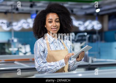 Junge Arbeitnehmerin in der Fischabteilung eines Ladens mit Tablette. Sie trägt eine Schürze und ein kariertes Hemd, was einen professionellen Einzelhandelsservice in einem modernen Ambiente veranschaulicht. Stockfoto