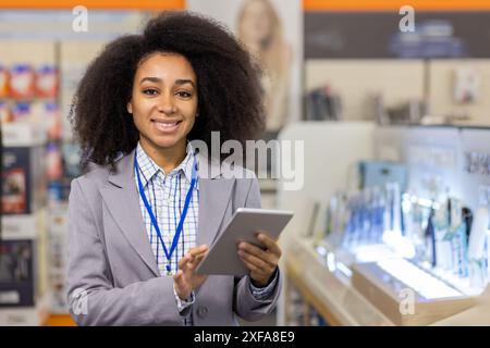 Ein lächelnder Mitarbeiter im Einzelhandel, der in einem Elektronikgeschäft steht und ein Tablet hält. Der Profi trägt einen Blazer und ein Namensschild, um Kunden beim Kauf von Technologie zu unterstützen. Stockfoto