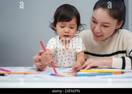 Kleinkind-Baby-Training zum Zeichnen mit Buntstift mit Mutter, die auf dem Tisch hilft Stockfoto