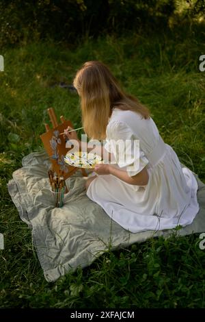 Ein schöner junger Künstler in einem weißen Kleid malt ein Bild am Flussufer Stockfoto