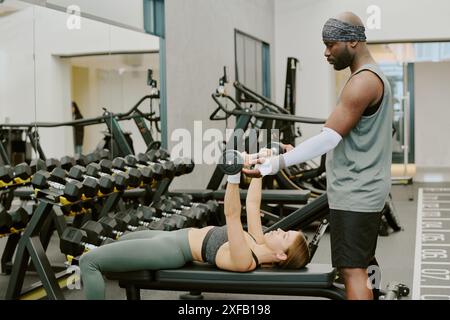 Afroamerikanischer Coach, der seiner Klientin mit sperrigen Muskeln hilft, sie drücken die Bank zusammen Stockfoto