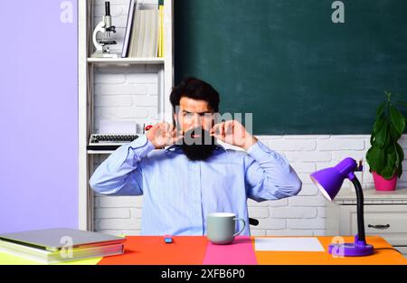 Ernsthafter Lehrer oder Schüler sitzt am Schreibtisch im Klassenzimmer. Schullehrer gibt Unterricht. Männlicher Professor in der Schulvorlesung. Ein bärtiger Mann, der an der universität studiert Stockfoto