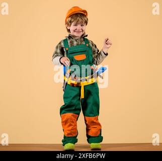 Lächelnder Junge in Baumeister-Uniform und Sicherheitshelm mit Werkzeuggürtel. Werkzeuge für das Bauen. Kleiner Reparaturmann mit Werkzeugen für die Reparatur. Süßes Kind in Overalls Stockfoto