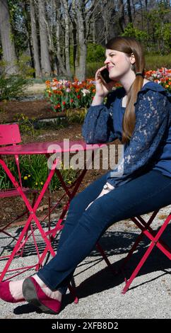 Die junge Frau sitzt an einem roten Metalltisch und -Stuhl. Sie ist in einem Garten und hat einen Anruf erhalten. Ihr Ausdruck ist unglücklich oder besorgt. Stockfoto