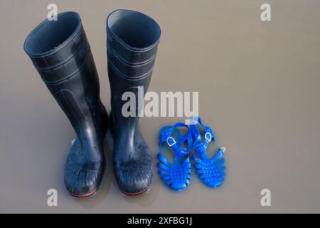 Schwarze Gummistiefel und blaue Kunststoffsandalen stehen in einer Regenpfütze am Strand. Es veranschaulicht die regnerischen Sommerferien und die extremen Wetterbedingungen, wi Stockfoto