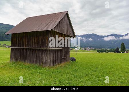 Alte Holzscheune steht auf einer breiten grünen Wiese mit einem Dorf im Hintergrund, gosau, alpen, österreich Stockfoto