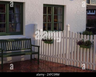 Gemütliche Gartenszene mit weiß gestrichenem Zaun und Bank neben einem Backsteinhaus mit grünen Fenstern, Greetsiel, Ostfriesland, Deutschland Stockfoto