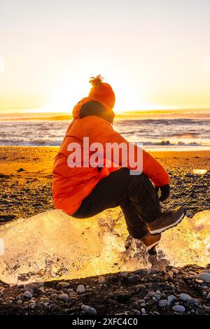 Ein Mann in einer orangefarbenen Jacke sitzt auf einem Felsen in der Nähe des Ozeans. Die Sonne untergeht und strahlt ein warmes Leuchten über die Szene. Der Mann genießt die Ruhe Stockfoto