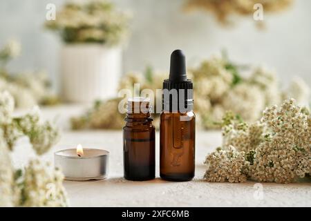 Flaschen mit ätherischem Aromatherapie-Öl mit Kerze und frischen Schafgarbenblüten auf einem weißen Tisch Stockfoto