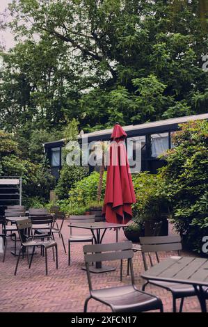 Ein Outdoor Cafe Sitzbereich mit leeren Stühlen und Tischen unter einem geschlossenen roten Sonnenschirm umgeben von Grün Stockfoto