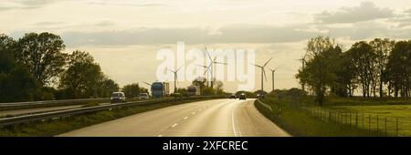 Eine asphaltierte Straße schlängelt sich durch eine ländliche Landschaft mit Bäumen an den Seiten und einer Linie von Windrädern in der Ferne vor einem klaren Sonnenuntergangshimmel. Stockfoto