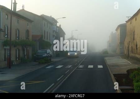 Ein einzelnes Auto fährt eine Straße in einem europäischen Dorf entlang, das in Morgennebel gehüllt ist. Die Gebäude entlang der Straße sind alt und charmant, mit Tradition Stockfoto