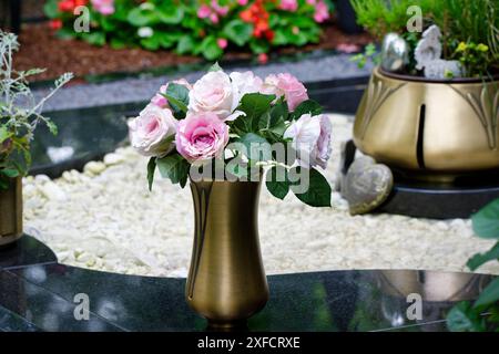 Eine schlanke Messingvase mit einem künstlichen Rosenstrauß auf einem Marmorgrab auf einem Friedhof Stockfoto