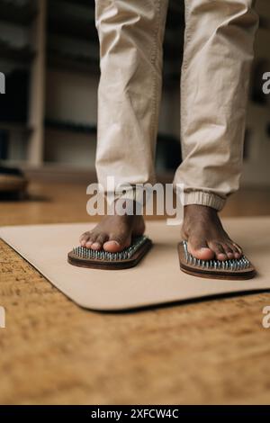 Vertikal geschnittene Aufnahme eines nicht erkennbaren afrikanischen Mannes, der auf einem hölzernen Yoga-Sadhu-Brett mit Metallnägeln steht, um zu Hause zu üben und zu meditieren. Stockfoto