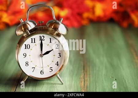 Stellen Sie Ihre Uhren zurück mit diesem Wecker mit farbenfrohen Herbstblättern über einem grünen rustikalen Holztisch. Sommerzeitkonzept. Stockfoto