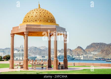 Goldene, verzierte Kuppelgazebo im traditionellen omanischen Stil mit Hafen und Kreuzfahrtschiffen im Hintergrund, Muttrah, Muscat, Oman Stockfoto