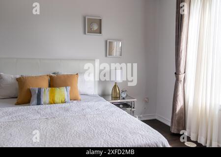 Hell beleuchtetes Schlafzimmer mit weißem Bettüberwurf, braunen und gelben Akzentkissen und Spiegeltisch. Aufgenommen in Toronto, Kanada. Stockfoto