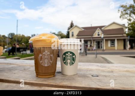 Zwei Starbucks-Drinks im Sonnenlicht - Holztisch im Freien - mit Karamell-Nieseln vereist - verschwommenes Gebäude und Baumhintergrund. Aufgenommen in Toronto, Kanada. Stockfoto