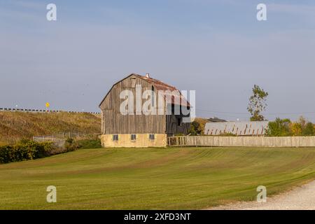 Rustikale Holzscheune - verwitterte Textur - vor klarem blauem Himmel - umgeben von üppigen grünen Feldern - entfernte Zaunlinie. Aufgenommen in Toronto, Kanada. Stockfoto