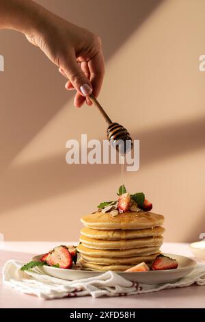 Frau gießt Honig auf Pfannkuchen mit Erdbeeren auf Rosa Stockfoto