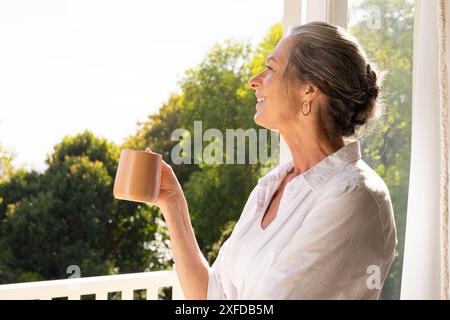 Lächelnde reife Frau, die Kaffeetasse hält und aus dem Fenster zu Hause blickt, Kopierraum Stockfoto