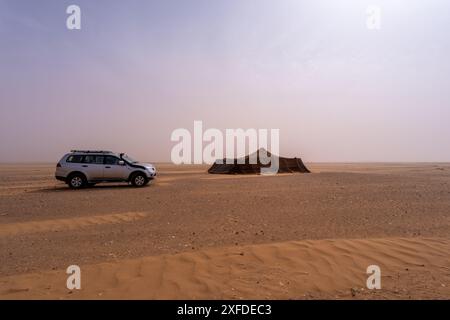 Ein Auto fährt während eines Staubsturms über die Sandebenen von Merzouga; die Sicht wird auf Dunst reduziert. Provisorische Unterkunft in Sicht, Marokko. Stockfoto