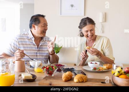 Genießen Sie das gemeinsame Frühstück, ein älteres Paar lacht und isst Croissants am Tisch Stockfoto