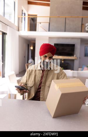 Paket empfangen, Mann im Turban, der Smartphone und Karton in der Küche hält Stockfoto