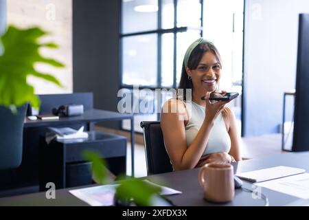 Lächelnde Frau im Büro, die ein Smartphone für Sprachnachrichten benutzt, am Schreibtisch sitzt Stockfoto