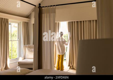 Vorhänge öffnen, ältere Frau in gelben Hosen genießen Morgensonnenlicht im gemütlichen Schlafzimmer, Kopierraum Stockfoto
