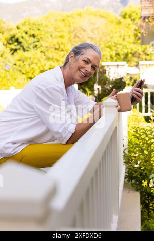 Lächelnde Seniorin mit Smartphone und Kaffeetasse, entspannend auf Balkongeländer Stockfoto