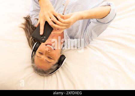 Musik mit Kopfhörern hören, Frau, die mit dem Smartphone auf dem Bett liegt Stockfoto